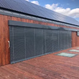 Drewniany budynek z panelami słonecznymi na dachu i szarymi osłonami okiennymi oraz drewnianym tarasem z wbudowanymi świetlikami