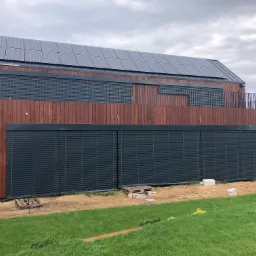 Nowoczesny dom z drewnianą elewacją i szarymi panelami słonecznymi na dachu, wyposażony w zewnętrzne systemy zaciemniające w kolorze antracytowym, częściowo opuszczone, widoczne palety i bloczki...