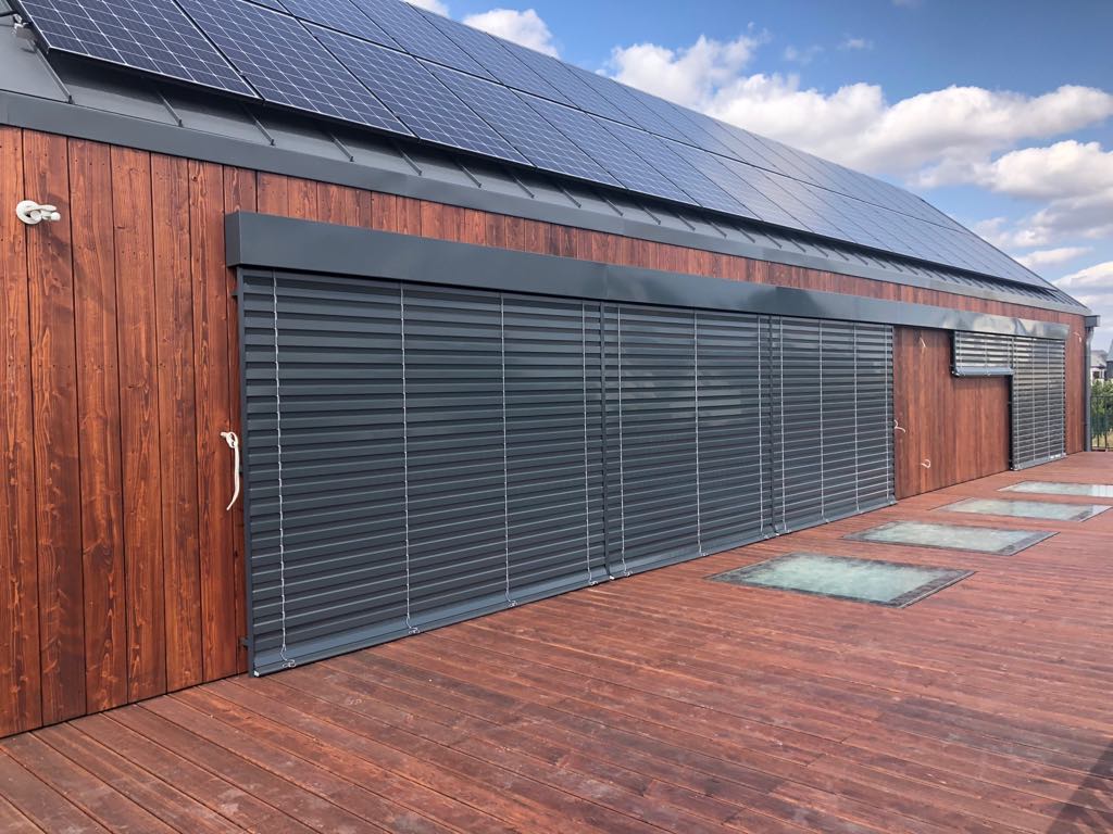 Drewniany budynek z panelami słonecznymi na dachu i szarymi osłonami okiennymi oraz drewnianym tarasem z wbudowanymi świetlikami