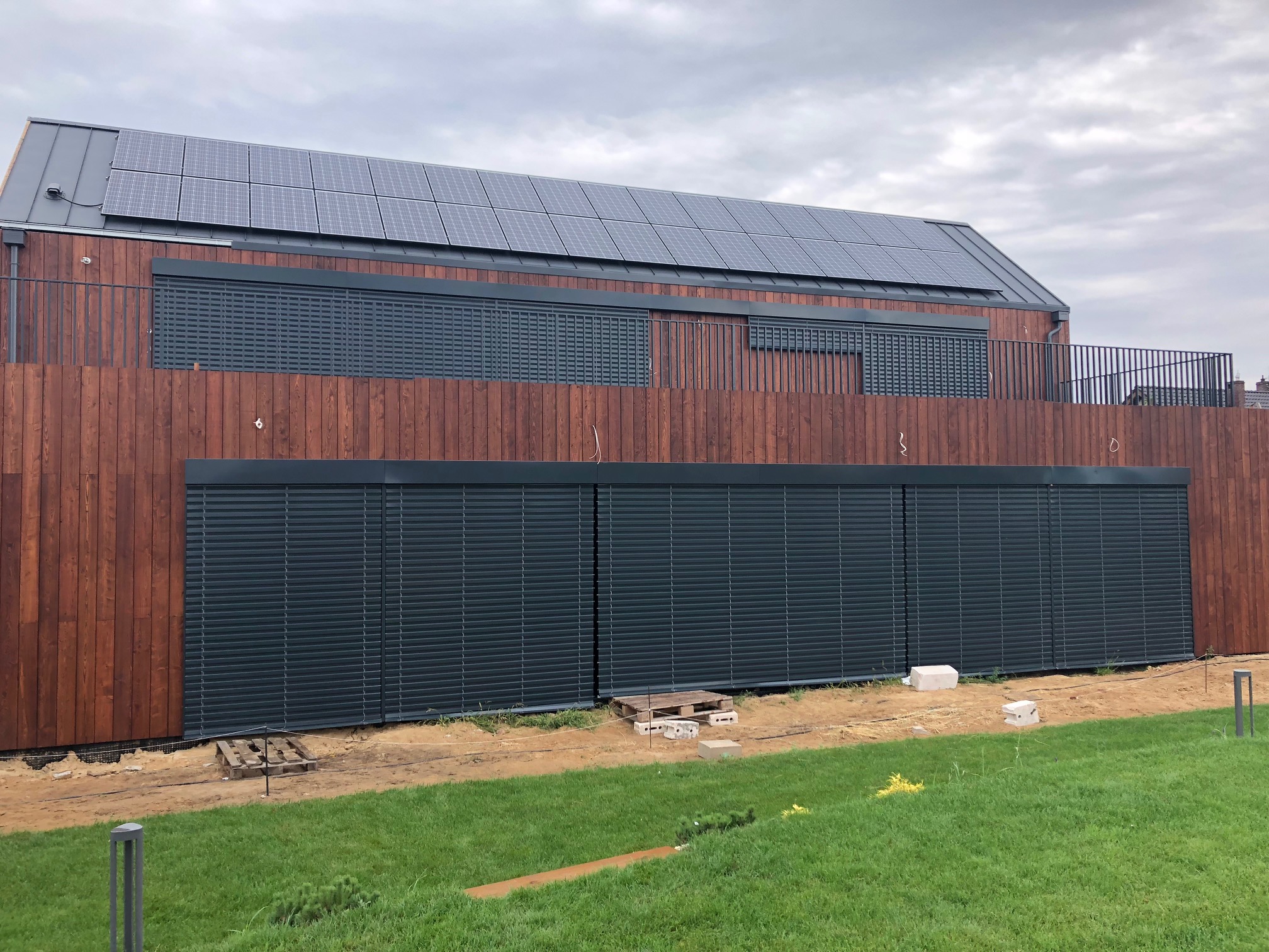 Nowoczesny dom z drewnianą elewacją i szarymi panelami słonecznymi na dachu, wyposażony w zewnętrzne systemy zaciemniające w kolorze antracytowym, częściowo opuszczone, widoczne palety i bloczki...