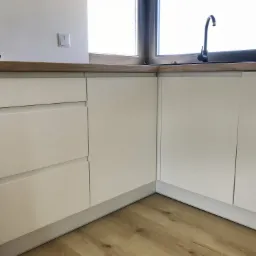 Fragment białej zabudowy kuchennej z drewnianym blatem, czarnym zlewem i baterią, widok z niskiego kąta na drewnianą podłogę.