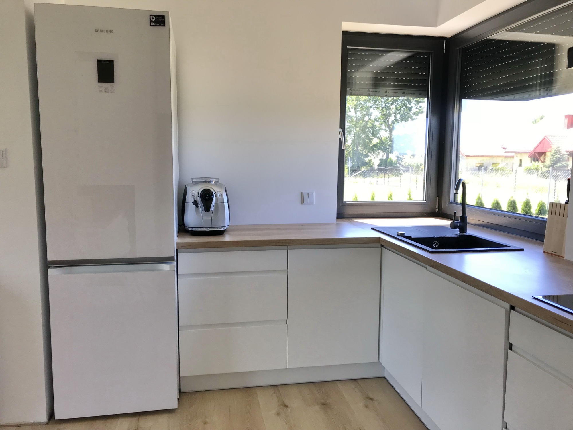 Nowoczesna kuchnia z białymi, matowymi frontami, drewnianym blatem i czarnym zlewem pod oknem z roletami zewnętrznymi, obok stoi biała lodówka Samsung i ekspres do kawy.
