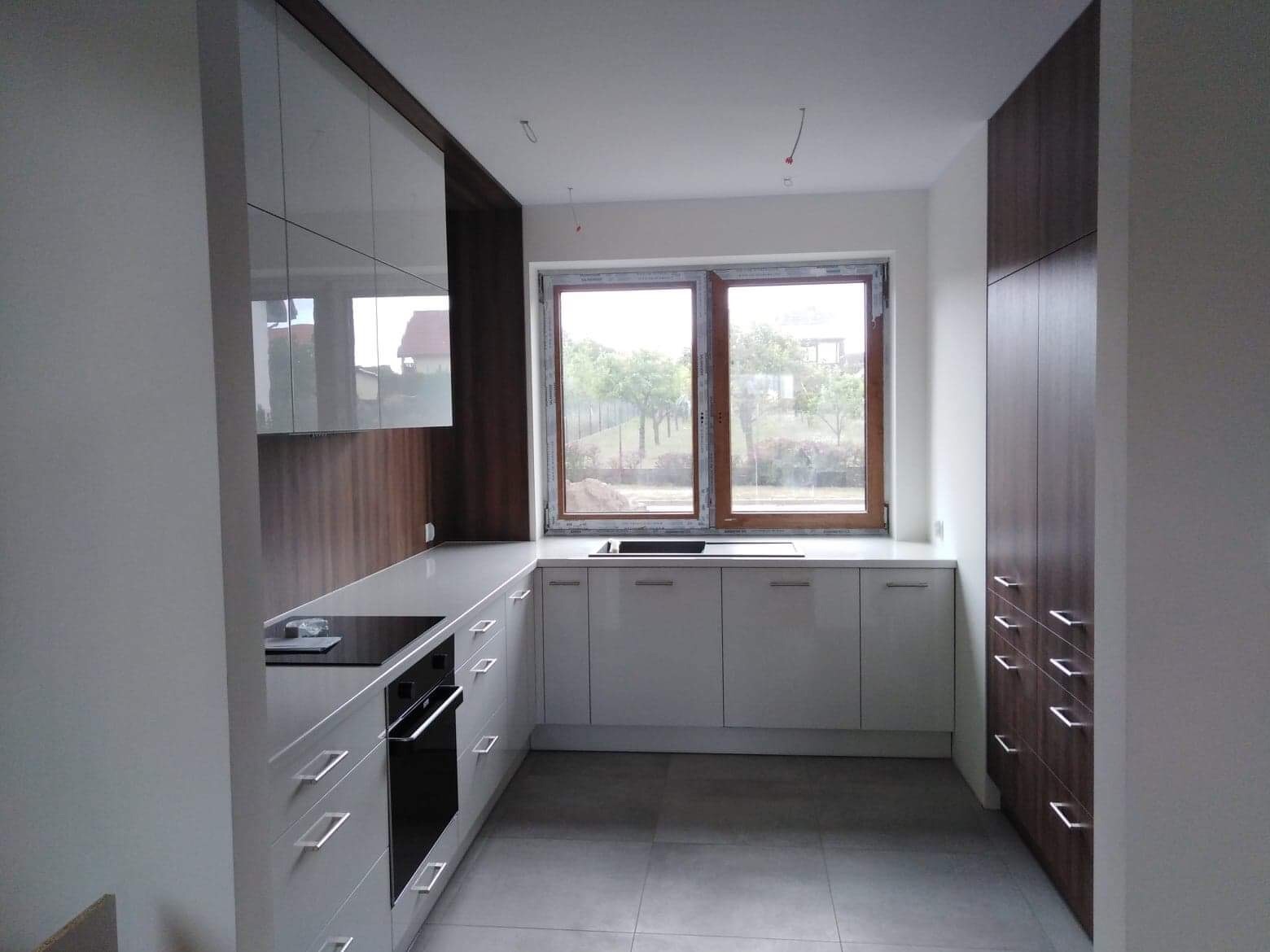 Nowoczesna kuchnia z białymi szafkami i drewnianymi akcentami, widok na ogród przez okno, szara podłoga.