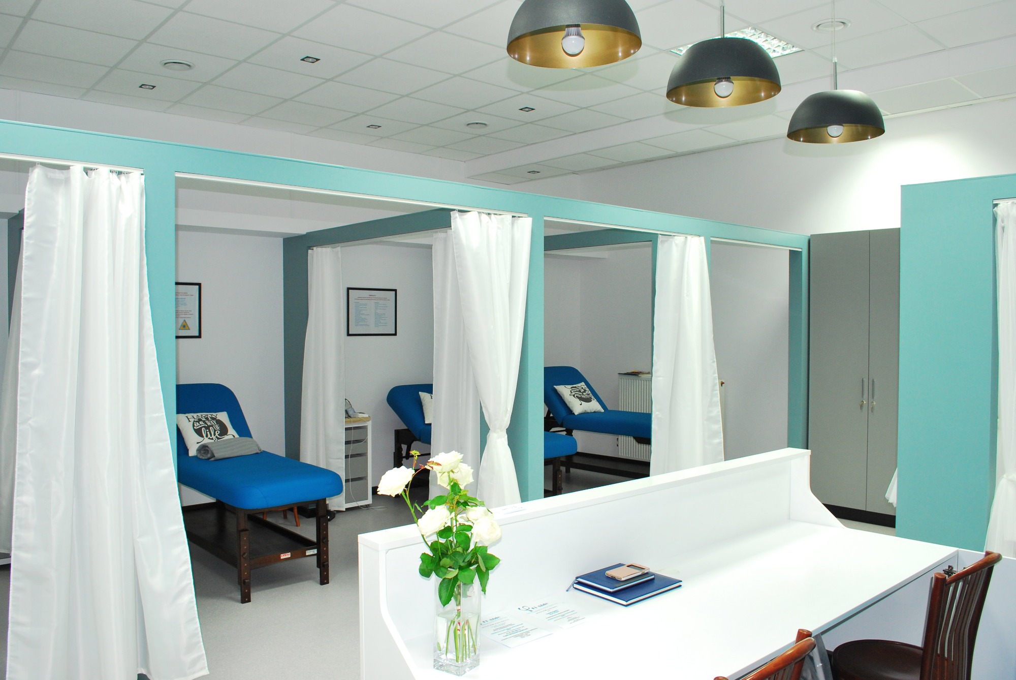 Jasne wnętrze gabinetu z trzema niebieskimi leżankami oddzielonymi białymi zasłonami, kwiaty w wazonie na białym biurku z notatnikami i telefonem, wiszące lampy z złotym wnętrzem.