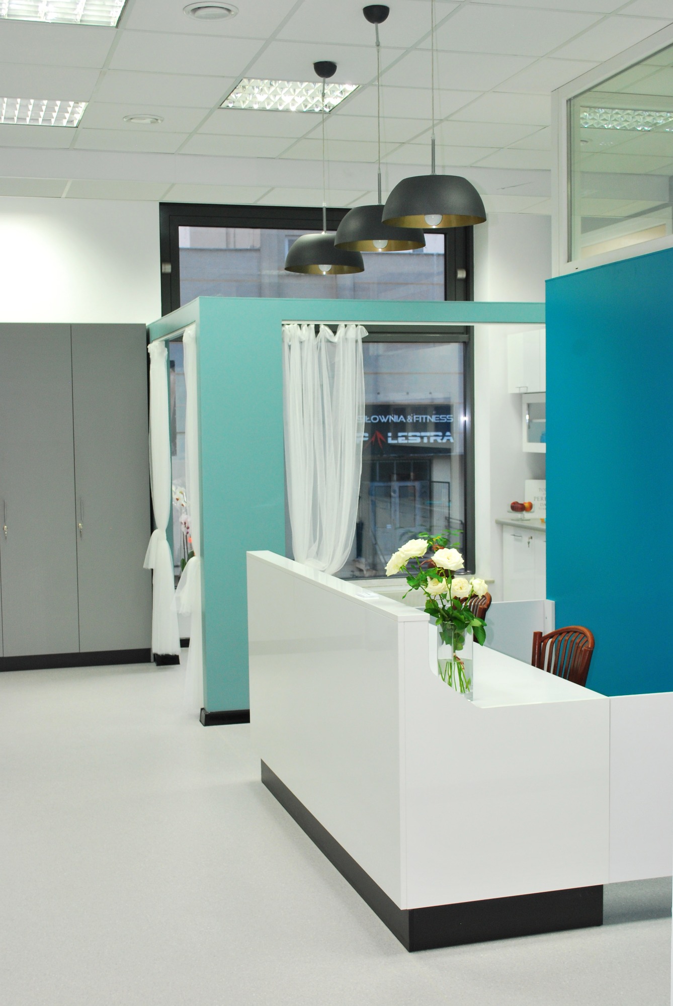 Biała lada recepcyjna z wazonem białych róż, trzy czarne lampy wiszące, szare szafy w tle, turkusowe ściany działowe i okno z widokiem na siłownię.