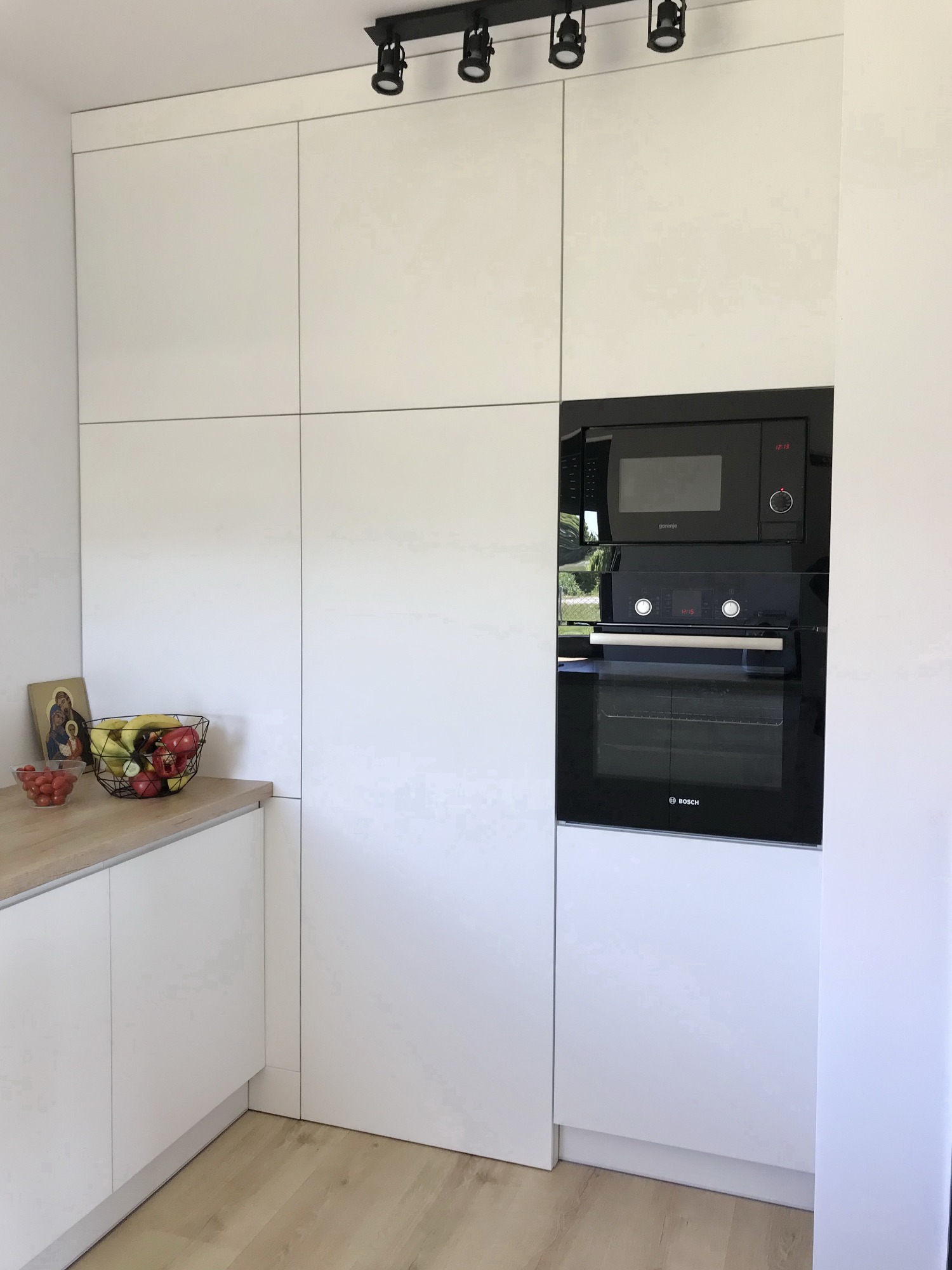 Biała zabudowa kuchenna z piekarnikiem i mikrofalówką w minimalistycznym stylu, na blacie koszyk z owocami i ikona religijna.