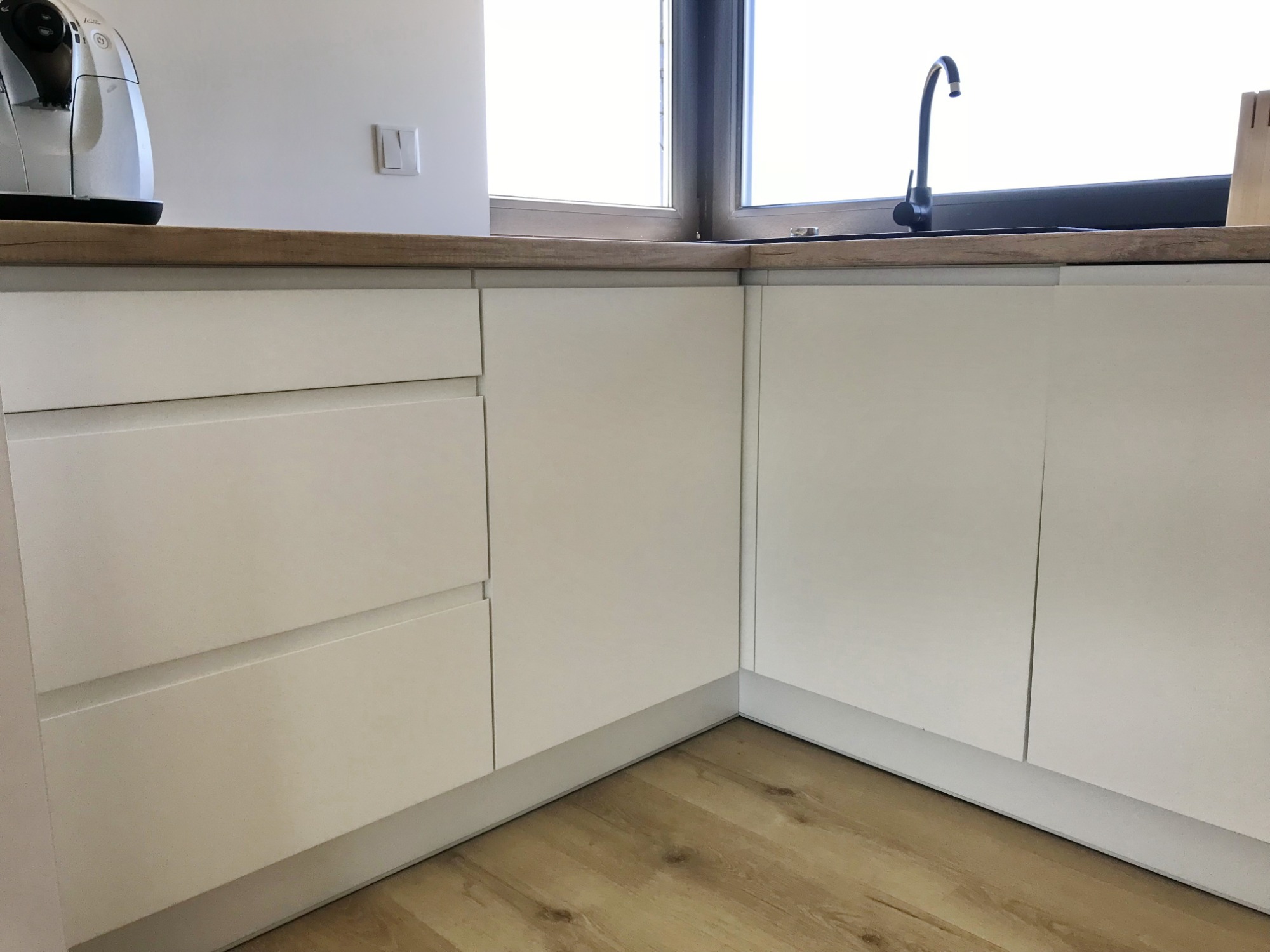 Fragment białej zabudowy kuchennej z drewnianym blatem, czarnym zlewem i baterią, widok z niskiego kąta na drewnianą podłogę.