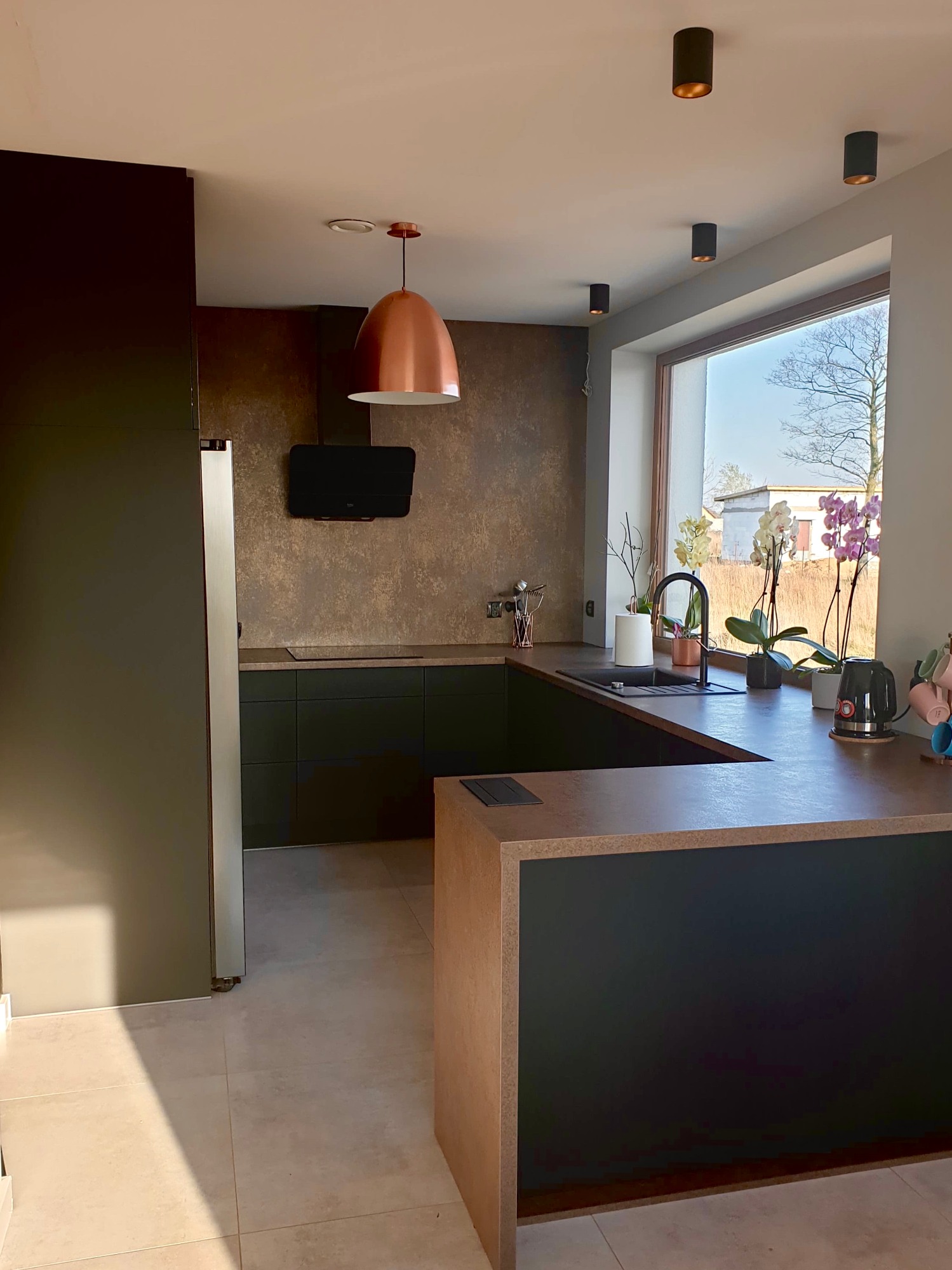 Nowoczesna kuchnia z ciemnozielonymi szafkami, drewnianym blatem i miedzianymi akcentami, w tym lampą wiszącą i detalami na ścianie, z widokiem na ogród przez duże okno.