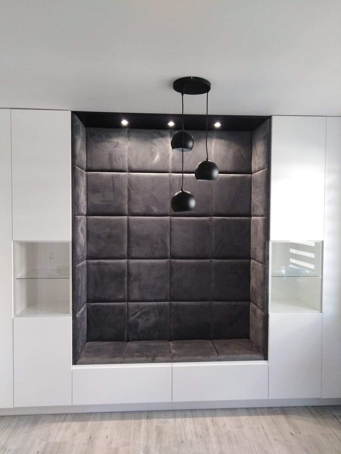 Biała szafa wnękowa z tapicerowanym, ciemnoszarym siedziskiem i oświetleniem punktowym oraz wiszącą lampą kulistą.
