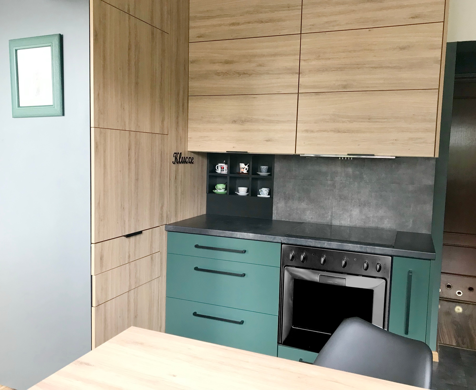 Nowoczesna kuchnia z drewnianymi szafkami górnymi i zielonymi dolnymi, czarnym blatem i piekarnikiem, z dekoracyjnym napisem 'Klucze' i półką na filiżanki.