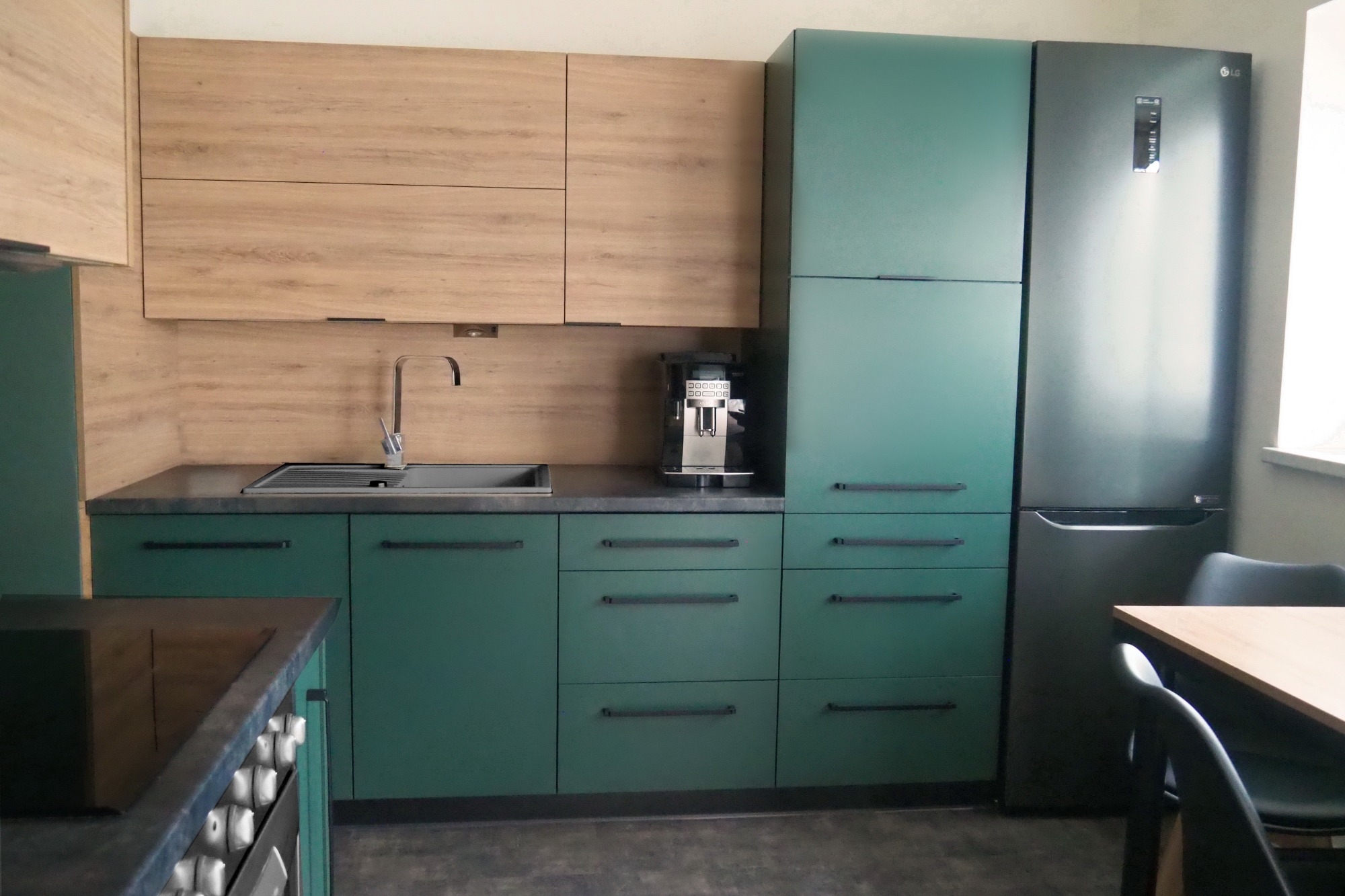 Nowoczesna kuchnia z zielonymi szafkami dolnymi i drewnopodobnymi górnymi, szarym blatem, srebrnym kranem i ekspresem do kawy.