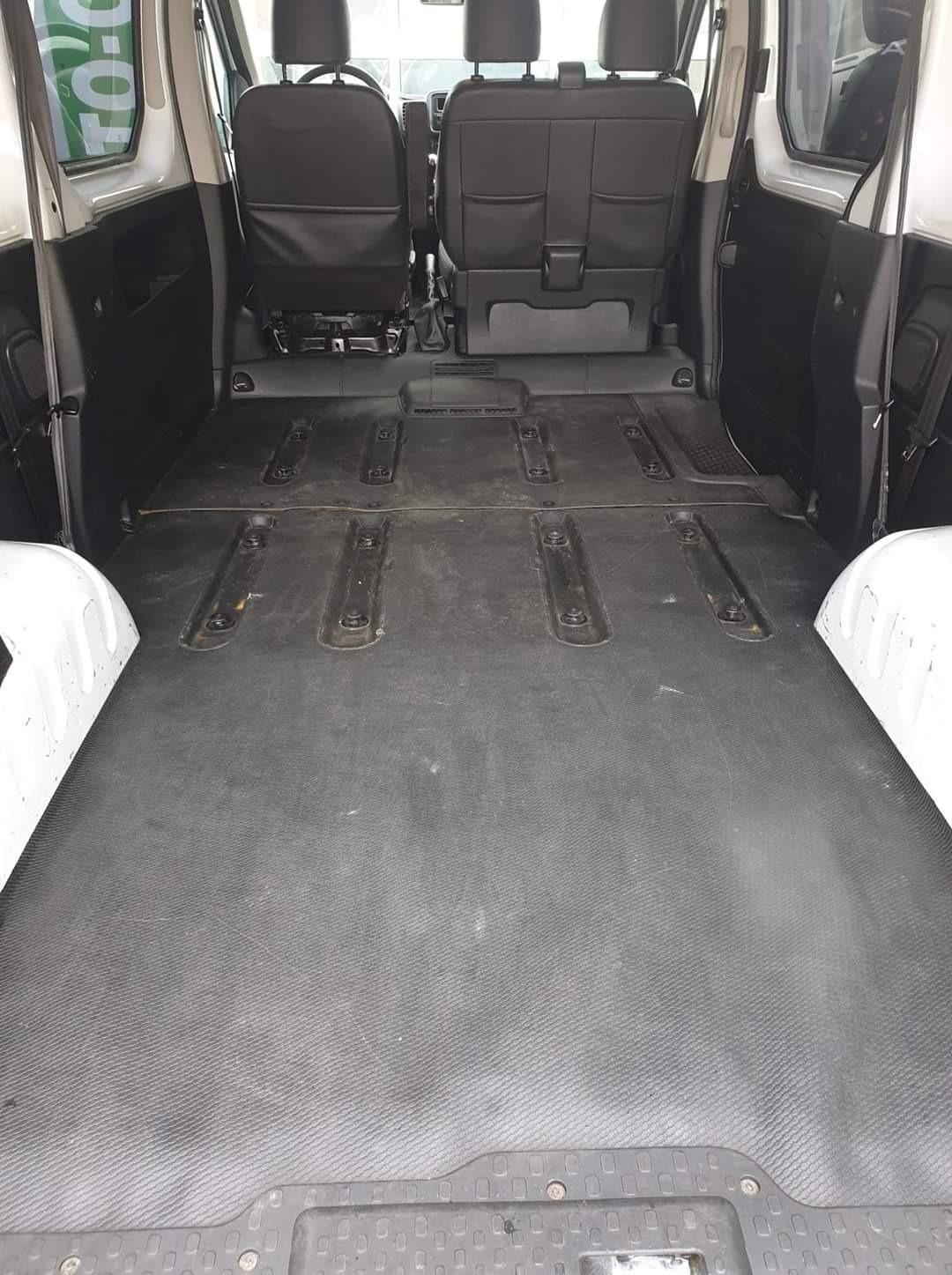 Pusta przestrzeń ładunkowa białego samochodu dostawczego z czarną gumową podłogą i zamontowanymi szynami mocującymi, widok od tyłu z widocznymi siedzeniami pasażerów.