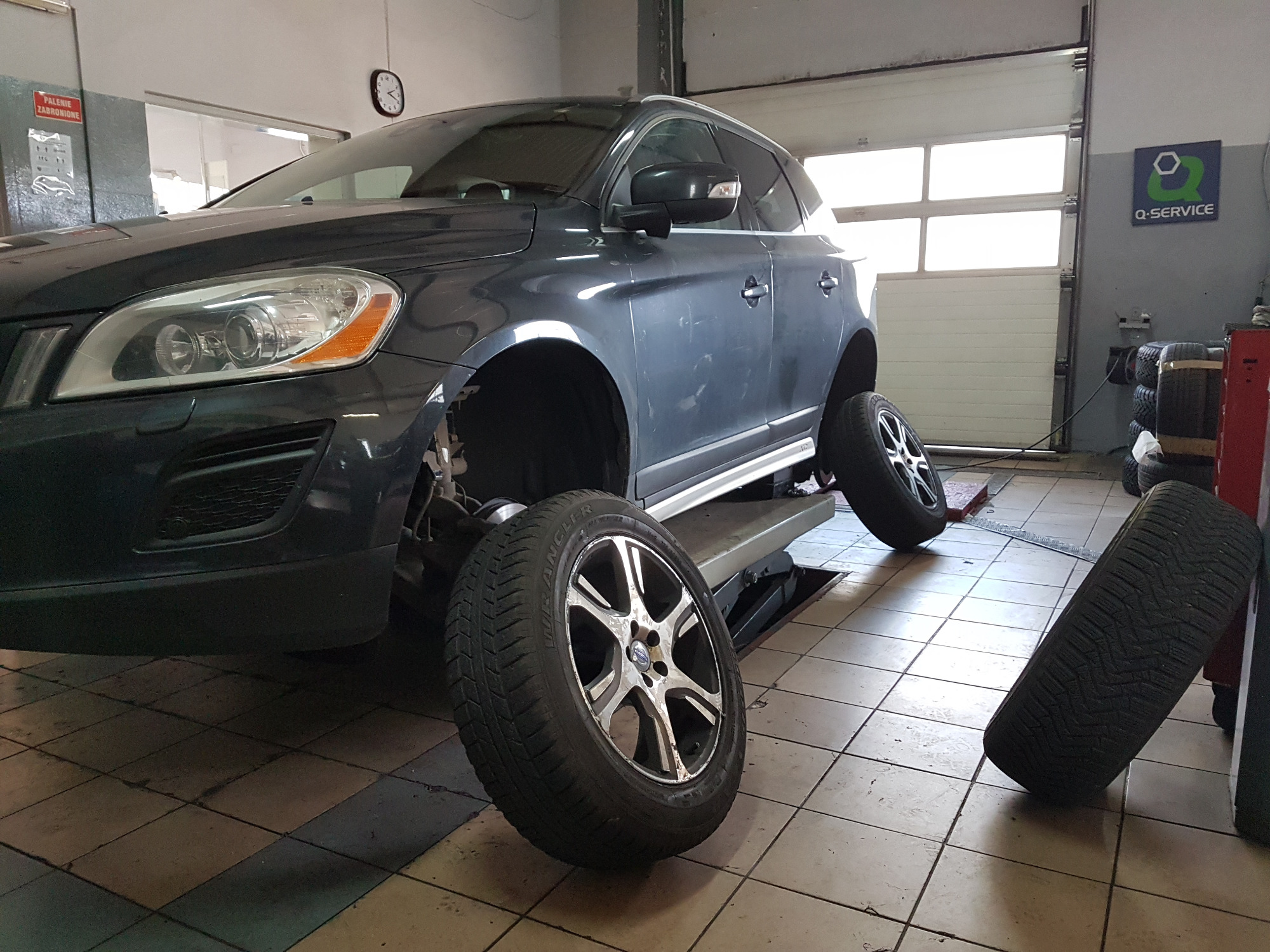 Szary samochód na podnośniku w warsztacie, zdjęte koła leżą obok, widoczny znak Q-Service.
