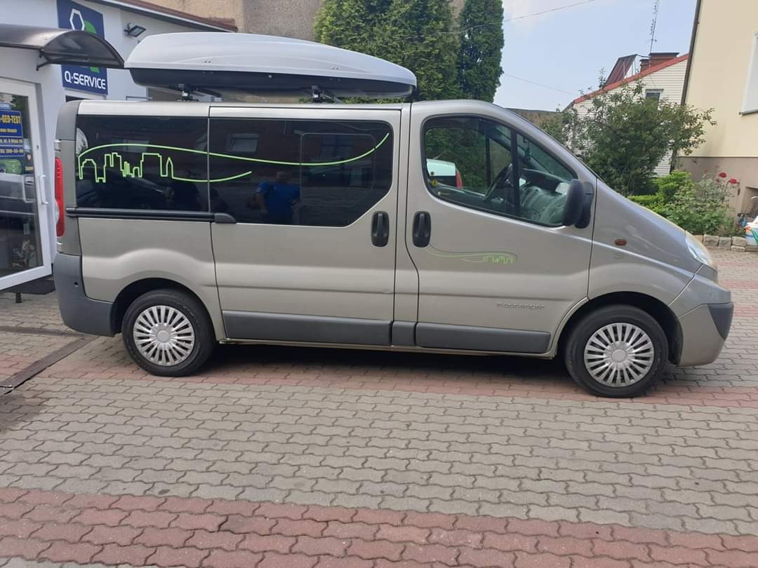Szary van Renault Trafic z bagażnikiem dachowym i zielonymi naklejkami przedstawiającymi panoramę miasta, zaparkowany na brukowanym placu przed budynkiem Q-Service.