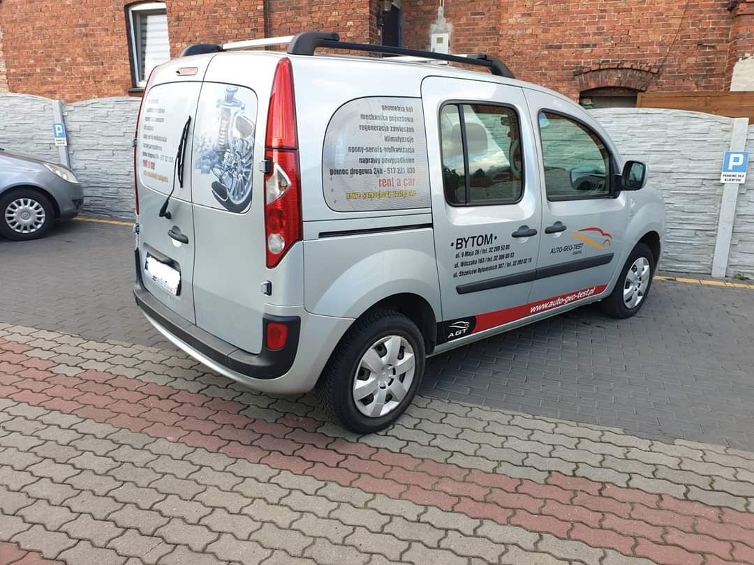 Srebrny samochód dostawczy z napisem 'rent a car' i adresem firmy AUTO-GEO-TEST zaparkowany na kostce brukowej przed ceglanym budynkiem. Na karoserii widoczne reklamy usług warsztatowych.