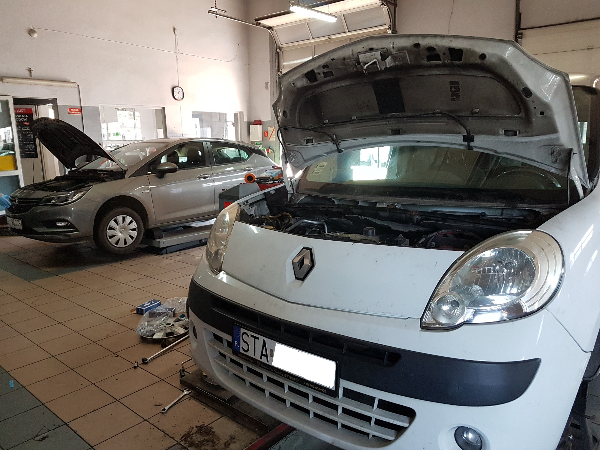 Dwa samochody z otwartymi maskami w warsztacie, jeden na podnośniku, narzędzia i części leżą na podłodze.