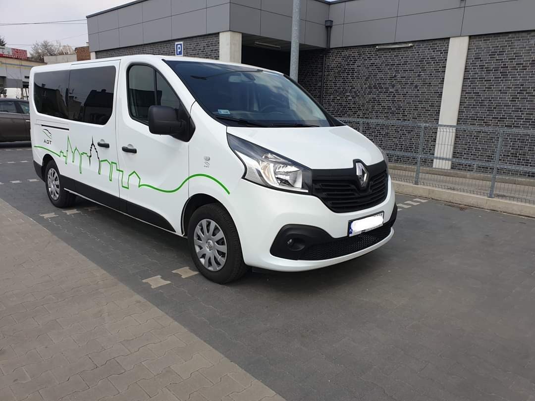 Biały Renault Trafic 9-osobowy z zielonym motywem miasta na boku, zaparkowany przed budynkiem z szarej cegły.