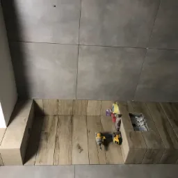 Fragment ściany z szarych, kwadratowych płytek ceramicznych i zabudowy z płytek imitujących drewno, z widocznymi narzędziami: dwoma wkrętarkami akumulatorowymi, taśmą malarską i akcesoriami...