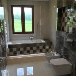 Nowoczesna łazienka z oknem na zieleń, marmurowe płytki w odcieniach szarości, biała wanna obudowana w szachownicę, wisząca toaleta i bidet.