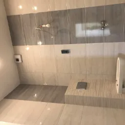 Nowa, jasna łazienka z szarymi akcentami: płytki w odcieniach beżu i szarości, wisząca toaleta ze spłuczką w ścianie, widoczne instalacje elektryczne i wodne.