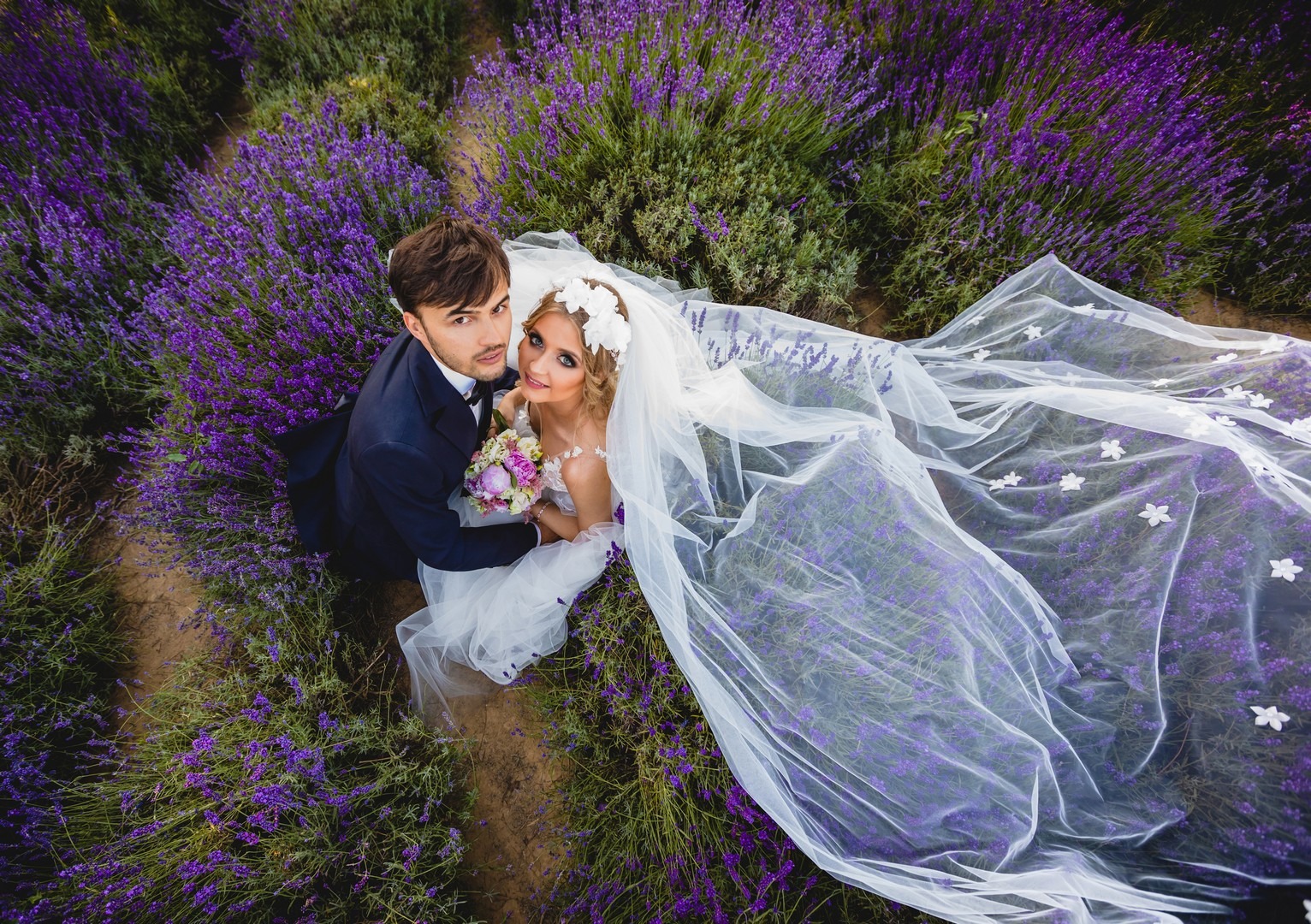 Para młoda w objęciach na polu lawendy, panna młoda w białej sukni i welonie z kwiatami, pan młody w granatowym garniturze, widok z góry.