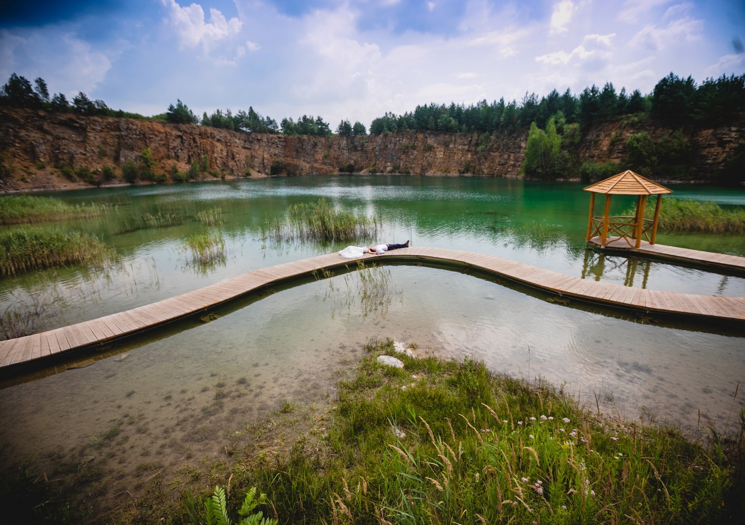 Para leży na drewnianym pomoście nad jeziorem w malowniczym kamieniołomie, widok z góry na nowożeńców i altankę.