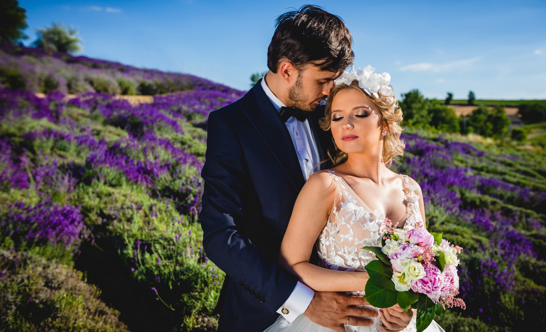 Para narzeczeńska w objęciach na tle pola lawendy, panna młoda trzyma bukiet kwiatów, pan młody w granatowym garniturze z muszką.