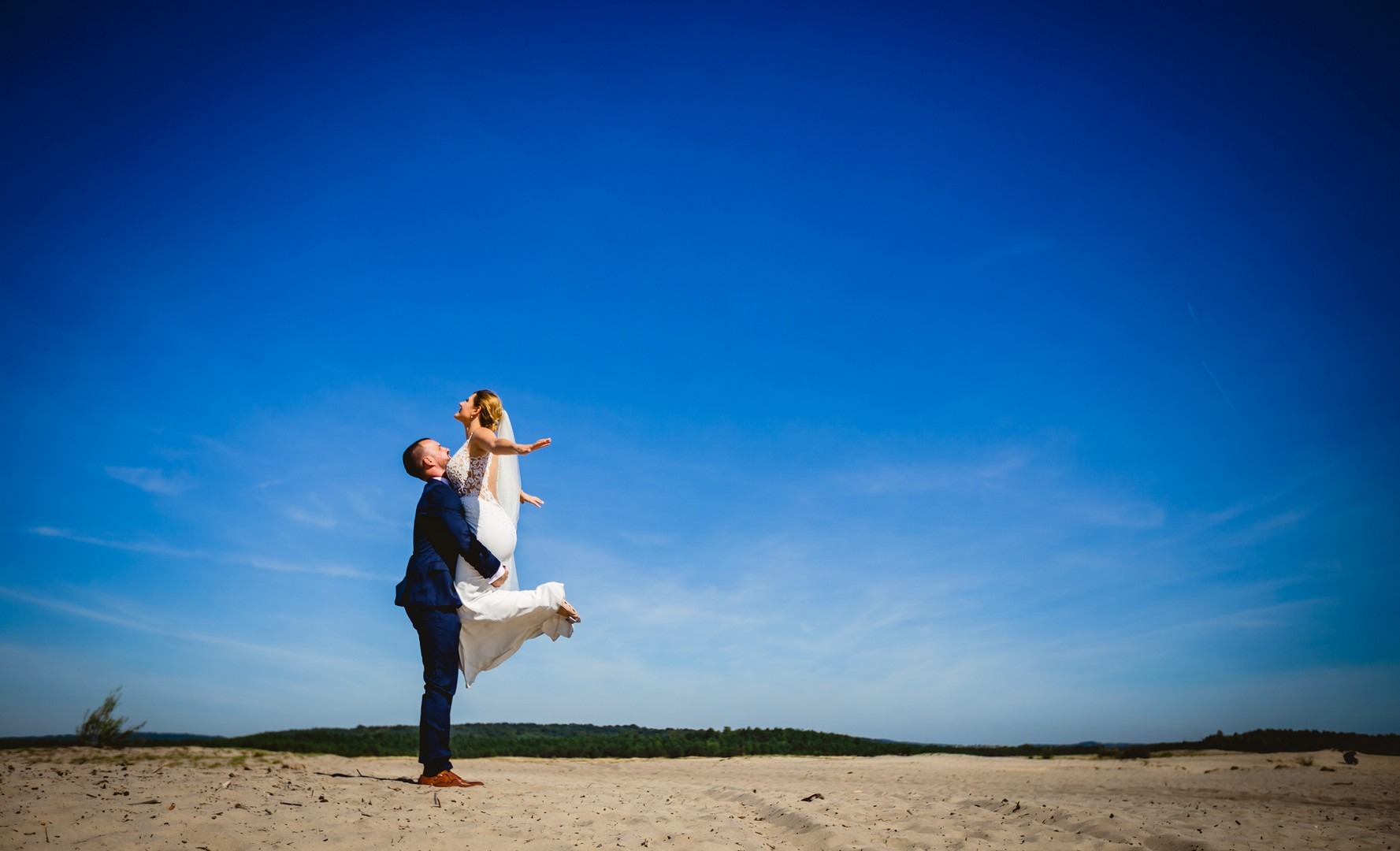 Para młoda na tle błękitnego nieba: pan młody w granatowym garniturze podnosi pannę młodą w białej sukni z koronkowymi detalami, na piaszczystym terenie z zielonymi drzewami w oddali.