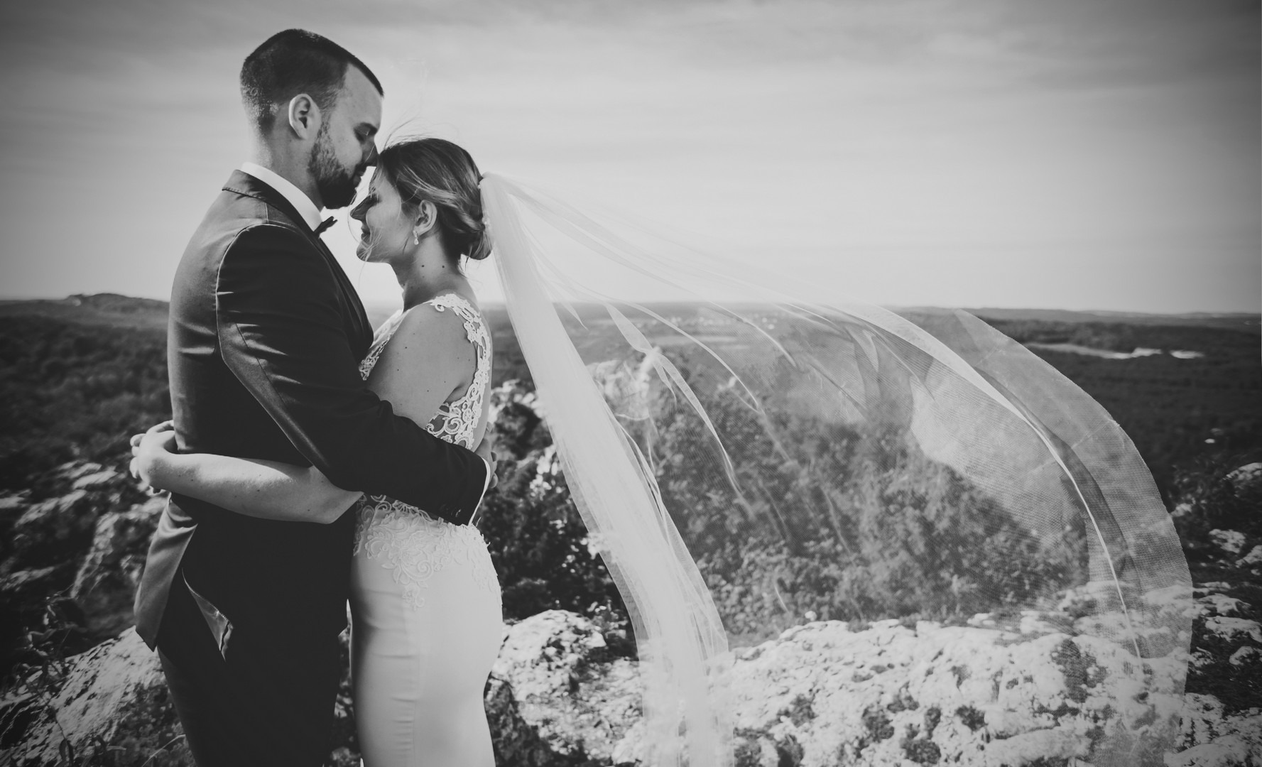 Czarno-białe zdjęcie pary młodej w objęciach na skalistym wzgórzu, welon powiewa na wietrze, ukazując odległy krajobraz.