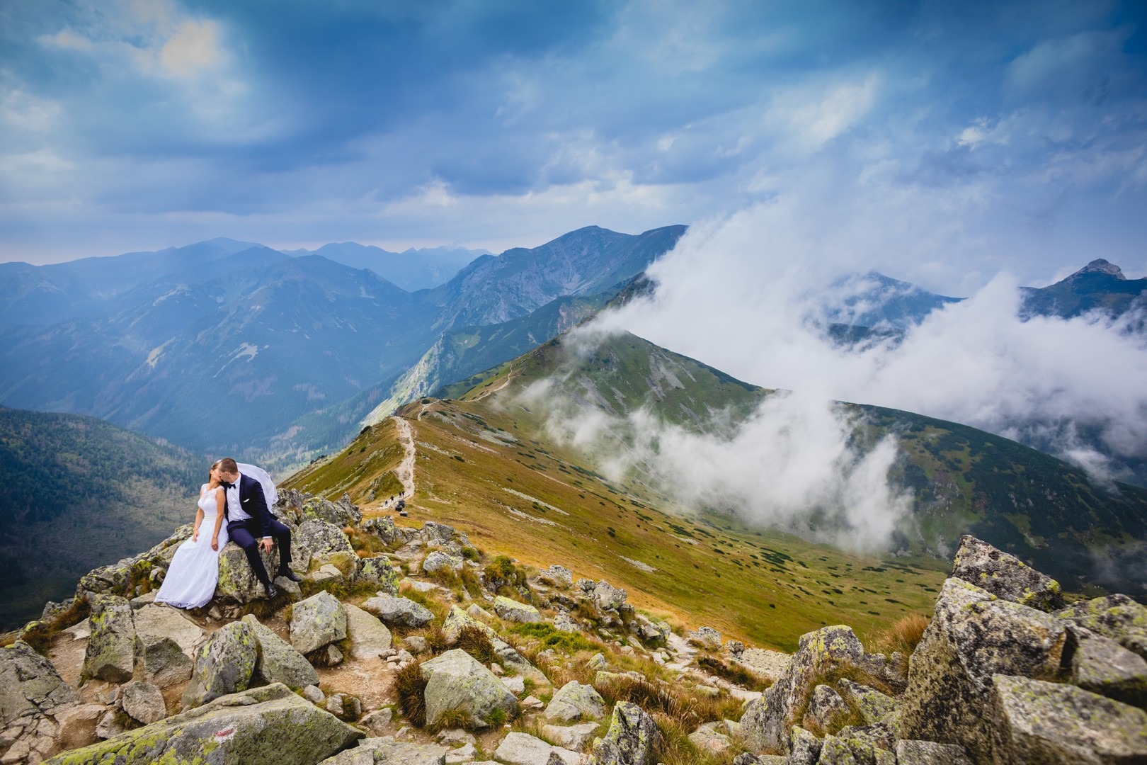 Para młoda w strojach ślubnych siedzi na kamieniach na szczycie góry, w tle widok na pasmo górskie spowite chmurami.