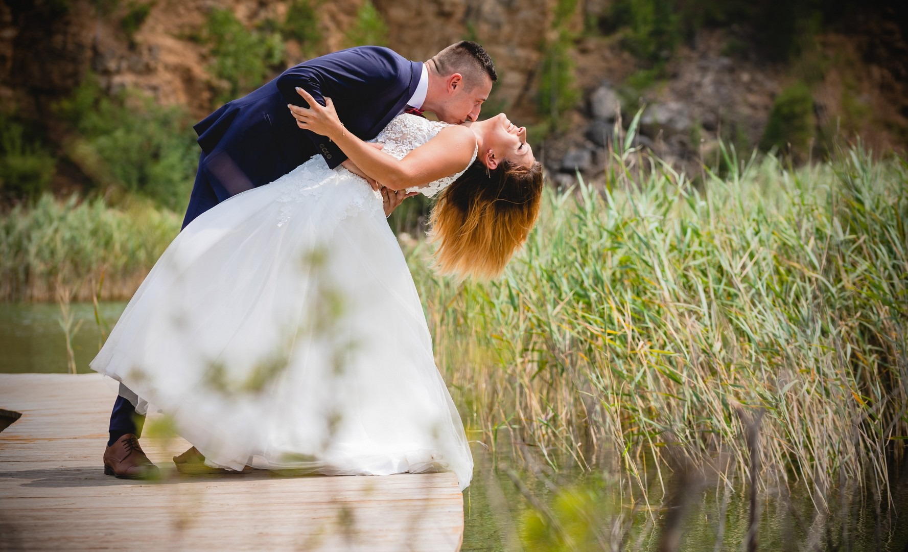 Para narzeczeńska w tańcu na drewnianym pomoście nad jeziorem, panna młoda w białej sukni z koronkowymi zdobieniami, pan młody w granatowym garniturze, w tle trzciny i skaliste zbocze.