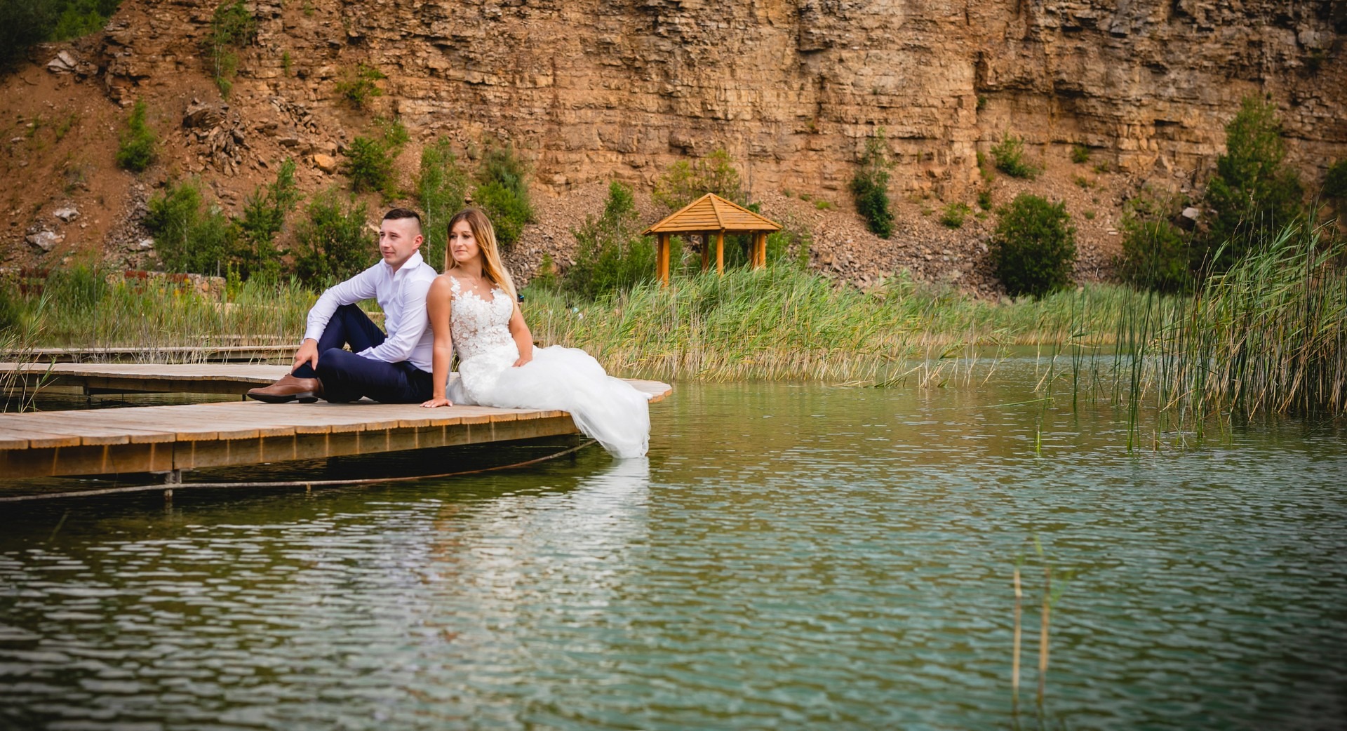 Para narzeczonych siedzi na drewnianym pomoście nad jeziorem, w tle skalna ściana i altanka.