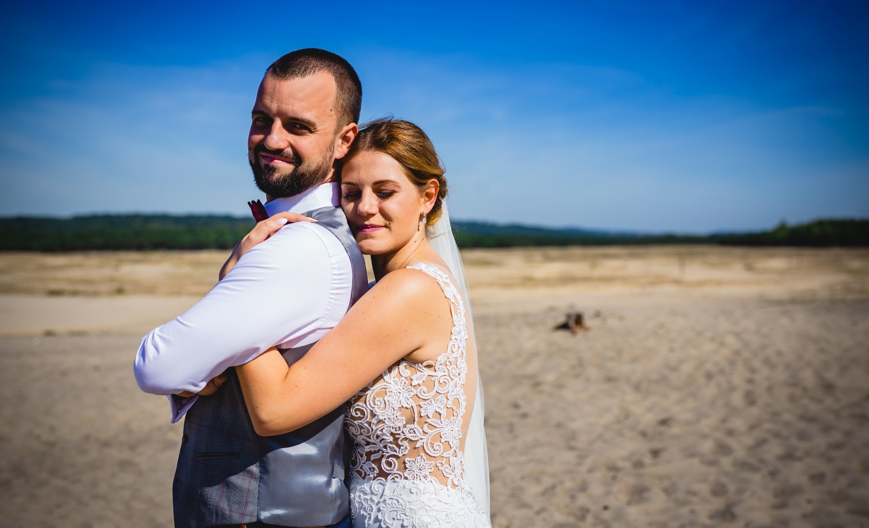 Para młoda w objęciach na piaszczystej scenerii w Mysłowie, panna młoda w koronkowej sukni, pan młody w kamizelce i muszce, błękitne niebo w tle.