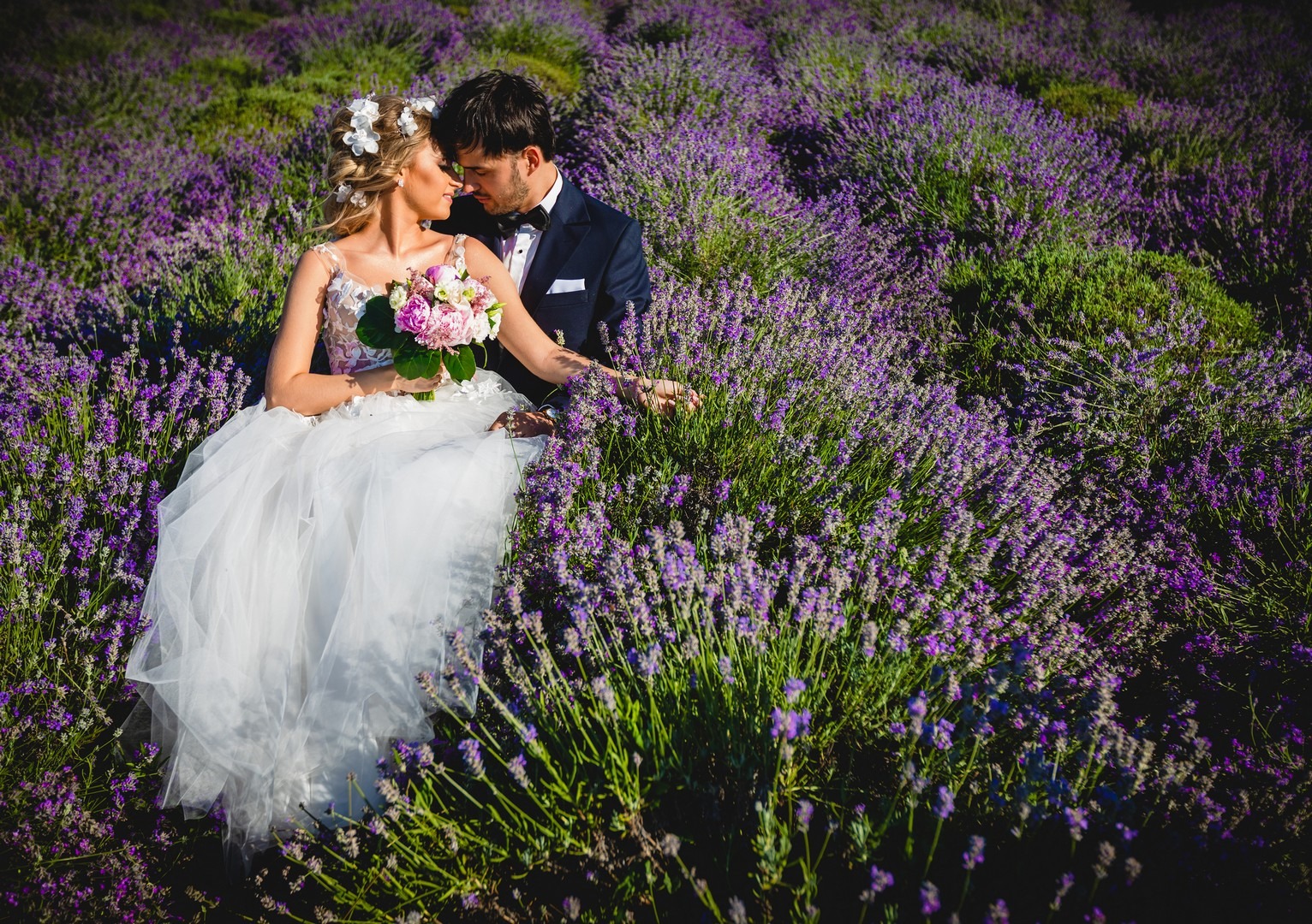 Para młoda w lawendowym polu, panna młoda w białej sukni trzyma bukiet różowych i białych kwiatów, pan młody w granatowym smokingu z muchą, delikatne dotykanie twarzami.