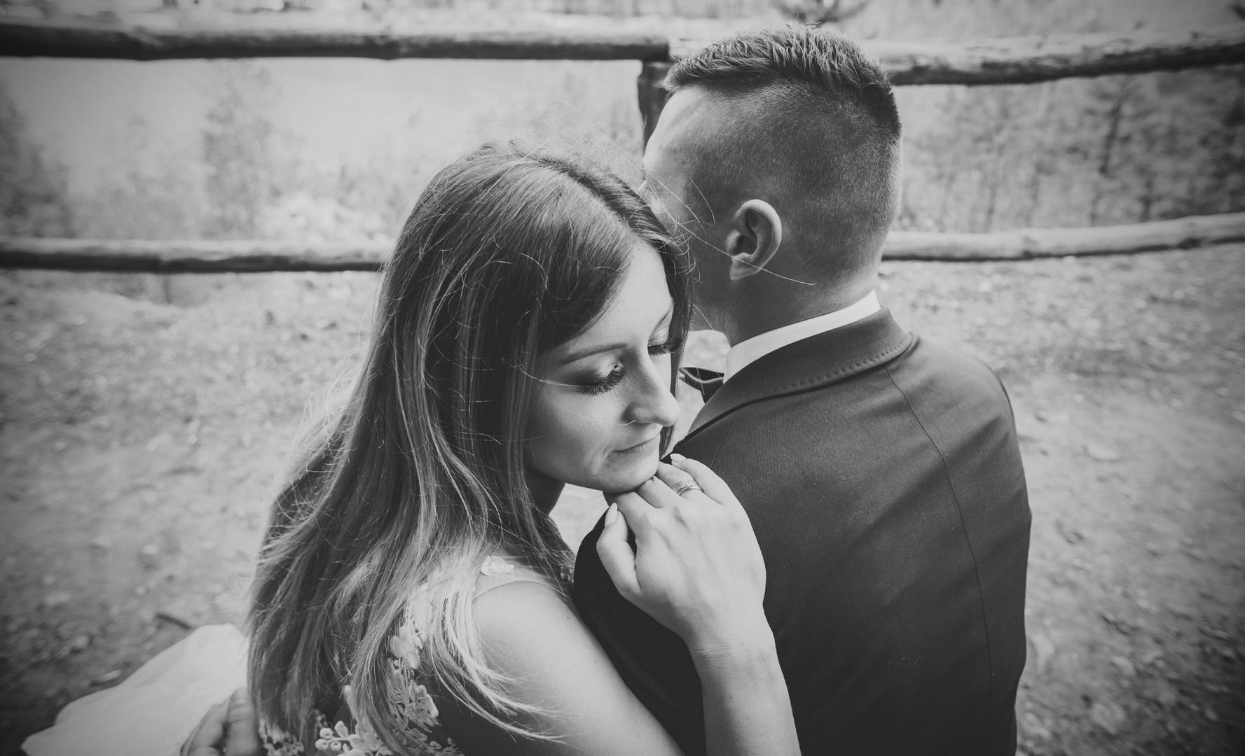 Czarno-białe ujęcie pary: panna młoda z długimi włosami opiera głowę o ramię pana młodego w garniturze, widoczny pierścionek zaręczynowy, tło z drewnianych bali.