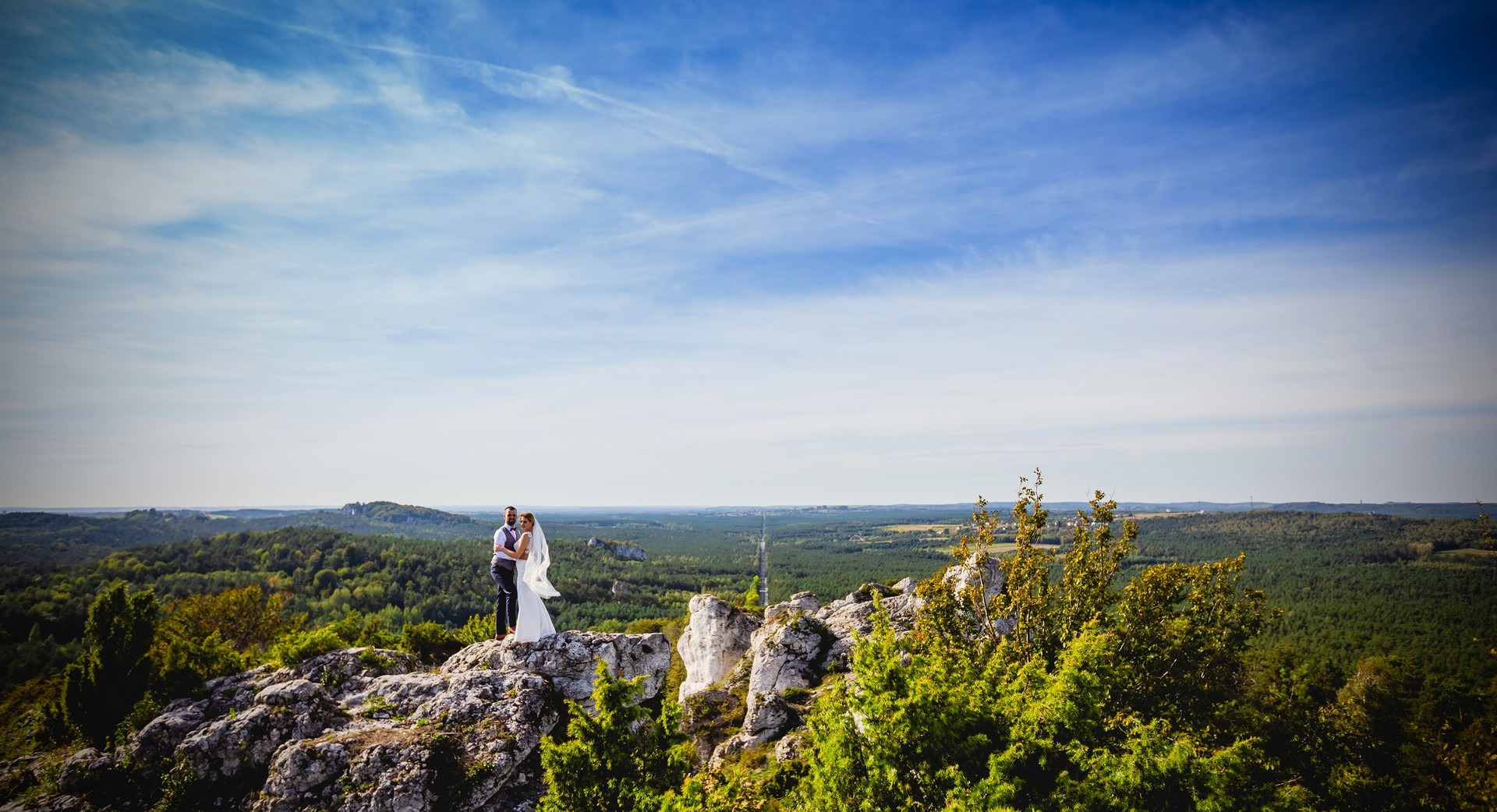 Para młoda w objęciach na skalistym szczycie z rozległą panoramą zalesionych wzgórz i błękitnym niebem z delikatnymi smugami chmur w tle.