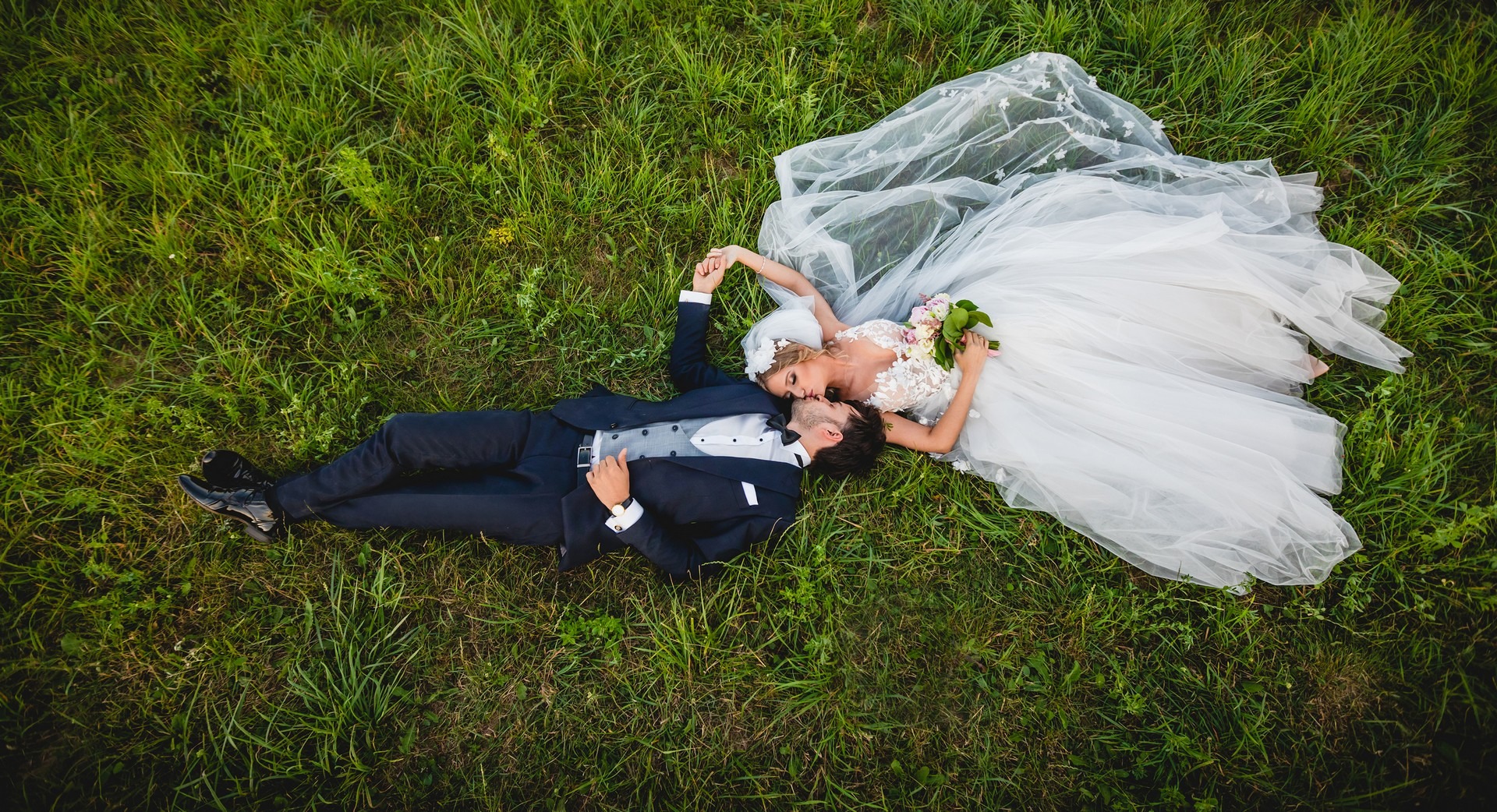 Para młoda w strojach ślubnych leży na zielonej trawie, trzymając się za ręce i całując się, widok z góry.