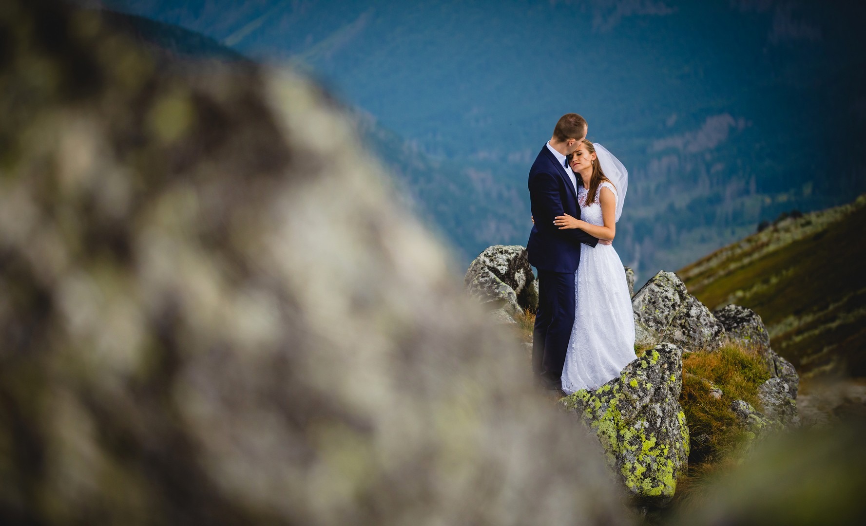 Para młoda w objęciach na szczycie skalistego wzgórza z widokiem na góry w tle, suknia ślubna z delikatnym welonem, garnitur w kolorze granatowym.