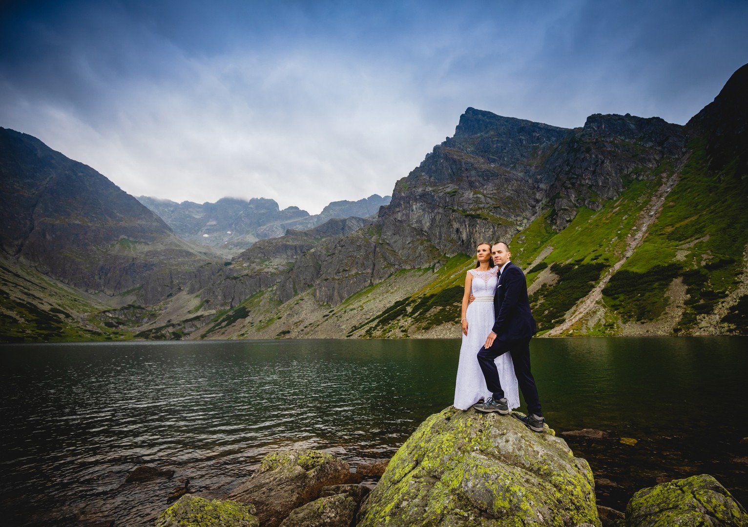Para młoda w strojach ślubnych pozuje na kamieniu porośniętym mchem nad brzegiem górskiego jeziora, w tle strome zbocza i szczyty Tatr.
