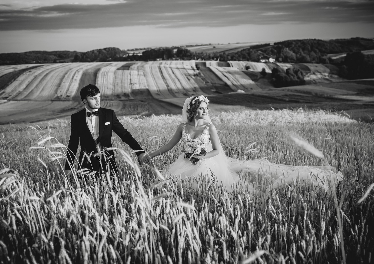 Czarno-białe zdjęcie pary młodej trzymającej się za ręce na polu zboża z pagórkowatym krajobrazem w tle.