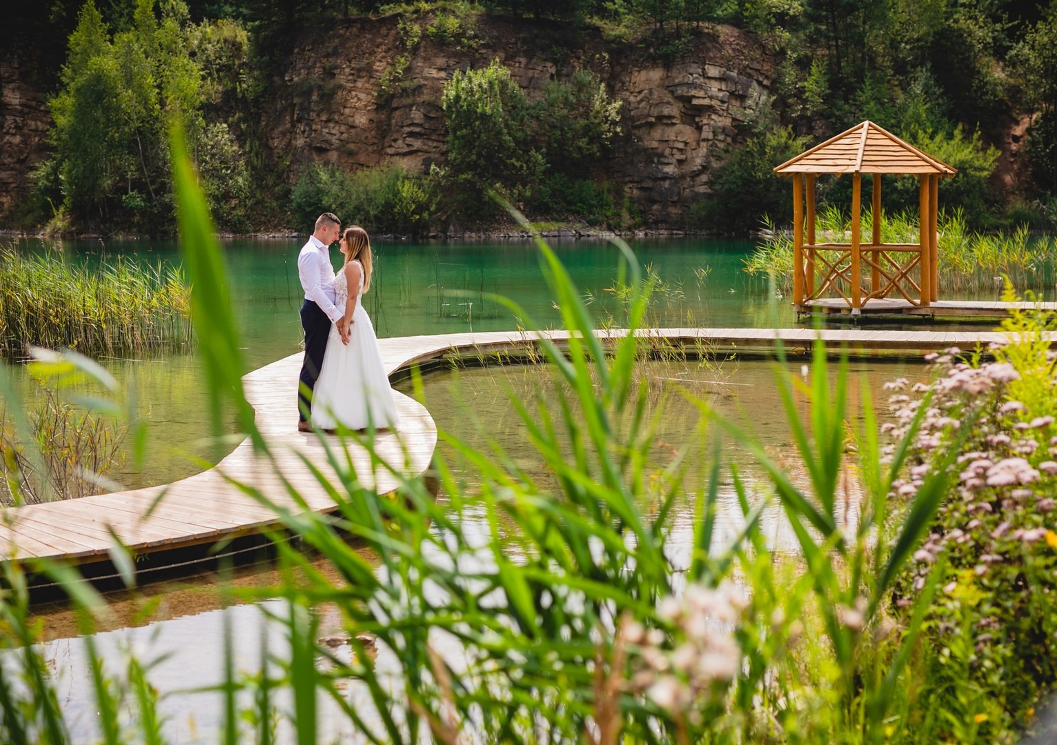 Para młoda w białej sukni i koszuli na drewnianym pomoście nad turkusową wodą, w tle skały, drzewa i drewniana altana
