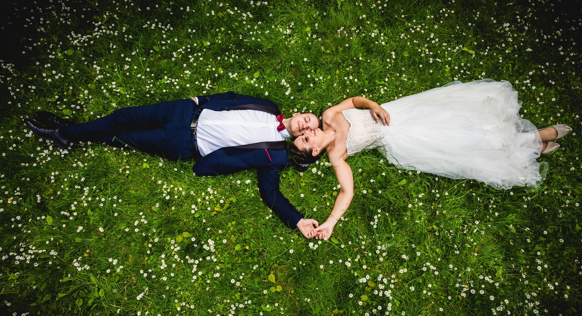 Para młoda leżąca na trawie pełnej stokrotek, widok z góry, ona w białej sukni ślubnej, on w granatowym garniturze z czerwoną muszką, trzymają się za ręce.