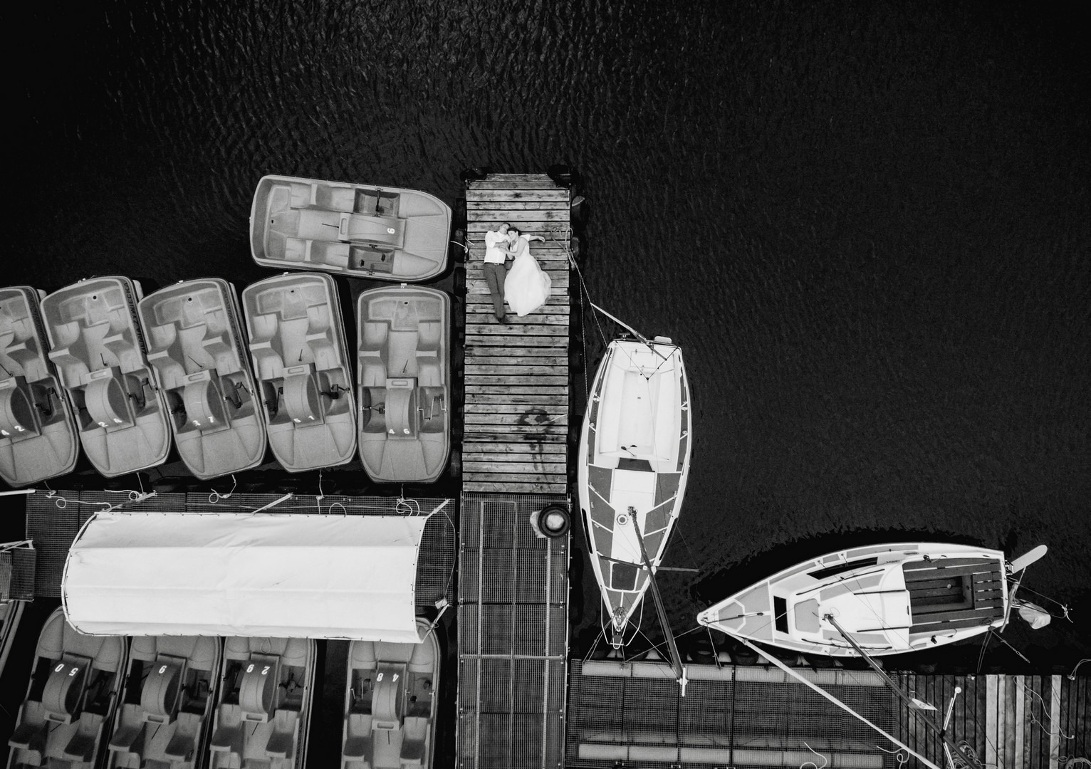 Zdjęcie z lotu ptaka: para młoda leżąca na drewnianym molo otoczonym łódkami i rowerami wodnymi na ciemnej wodzie.