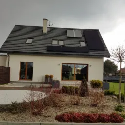 Nowoczesny dom jednorodzinny z panelami słonecznymi na dachu, widoczne okna dachowe, elewacja w jasnym kolorze, zadbany ogród z krzewami i trawą, pochmurne niebo.