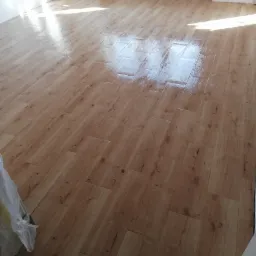 Świeżo ułożone, błyszczące panele podłogowe imitujące drewno w jasnym, pustym pomieszczeniu z białymi ścianami, widoczne nierówności na ścianach i brak listew przypodłogowych.