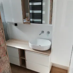 Nowoczesna umywalka nablatowa w owalnym kształcie, zamontowana na szafce z szufladami i półkami, widoczny fragment ściany z odsłoniętą cegłą, lustro z czarnym grzejnikiem drabinkowym w tle.