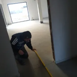 Pracownik w czapce i kamizelce klęczy, mierząc poziom podłogi żółtą poziomicą w surowym wnętrzu z widocznym oknem i ścianami w trakcie wykończenia.