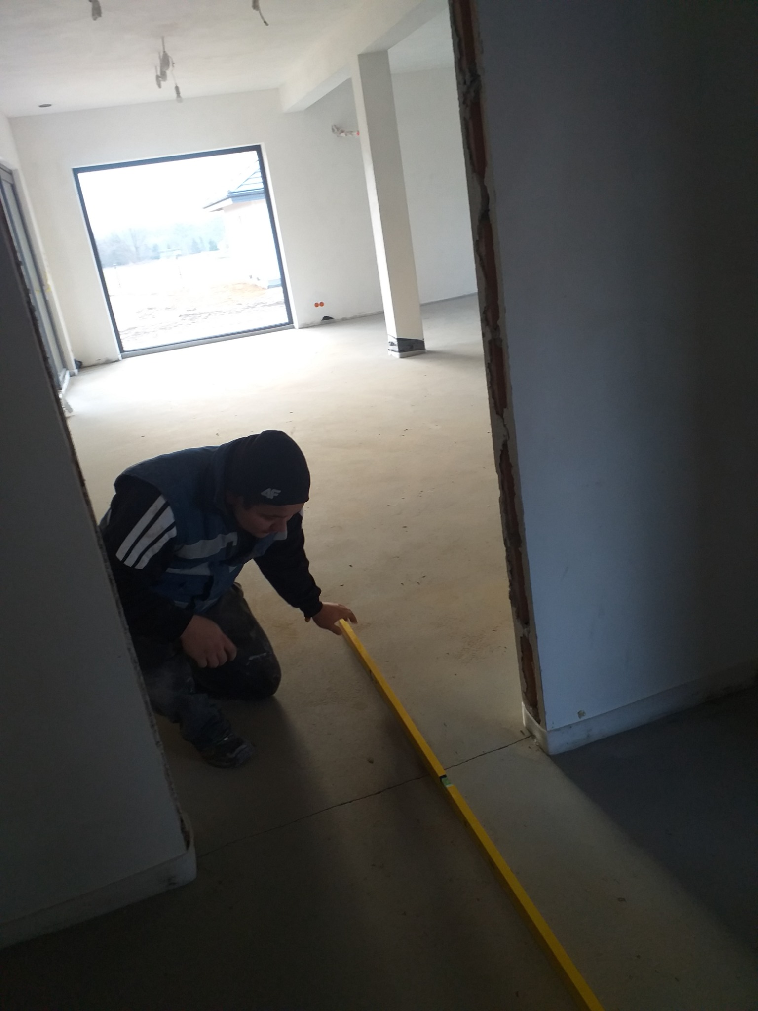 Pracownik w czapce i kamizelce klęczy, mierząc poziom podłogi żółtą poziomicą w surowym wnętrzu z widocznym oknem i ścianami w trakcie wykończenia.