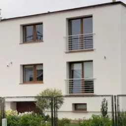 Dwupiętrowy dom z białymi ścianami, brązowymi ramami okiennymi i balkonami z metalowymi barierkami, widziany zza ogrodzenia z siatki, otoczony zielenią.