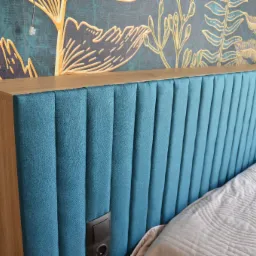 Wezgłowie łóżka tapicerowane na niebiesko z pionowymi przeszyciami, drewniana rama, gniazdko elektryczne, na tle tapeta z motywem roślinnym.